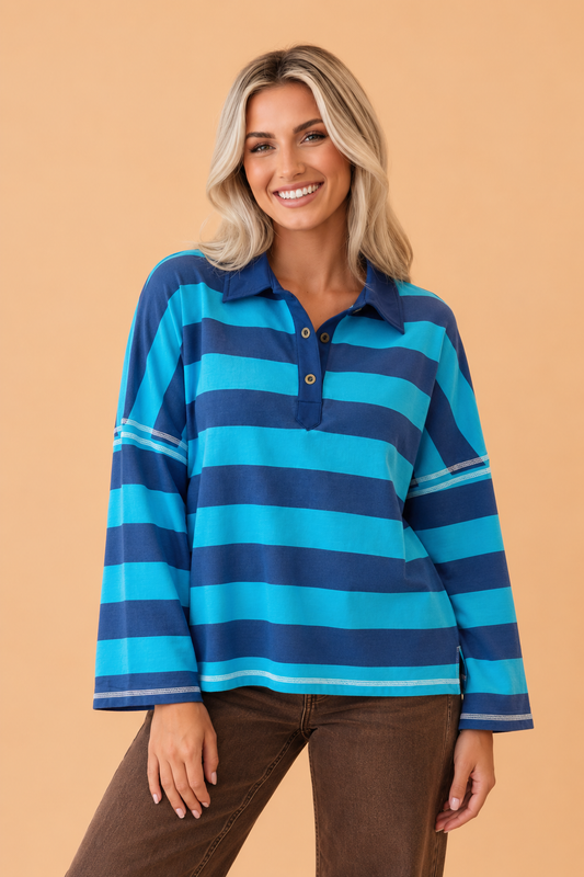 A smiling blonde woman wearing a Pretty Bash long-sleeve polo shirt with horizontal stripes in turquoise, medium blue, and navy, paired with brown pants, set against a solid peach studio background.