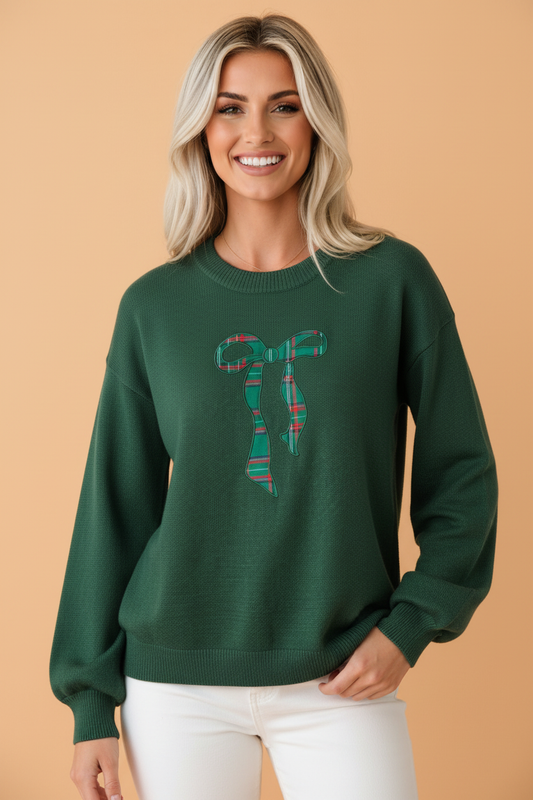 A smiling blonde woman wearing PRETTY BASH a dark green crewneck sweater with a plaid bow graphic on the chest, paired with white pants, set against a solid peach studio background.